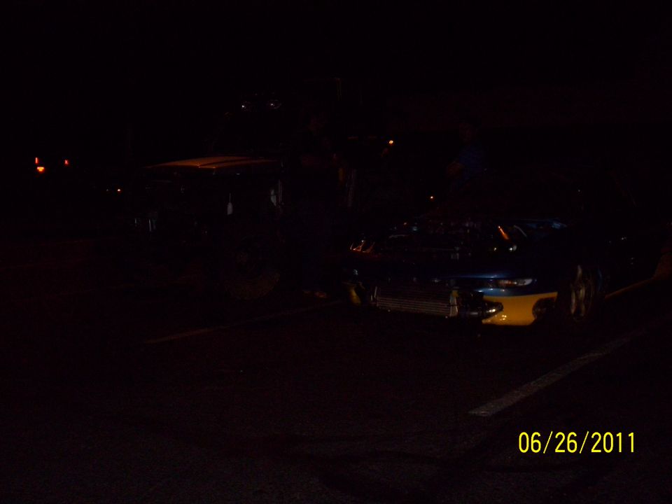 Front of a dark colored car with yellow accents, parked at night. Another vehicle is visible to the left