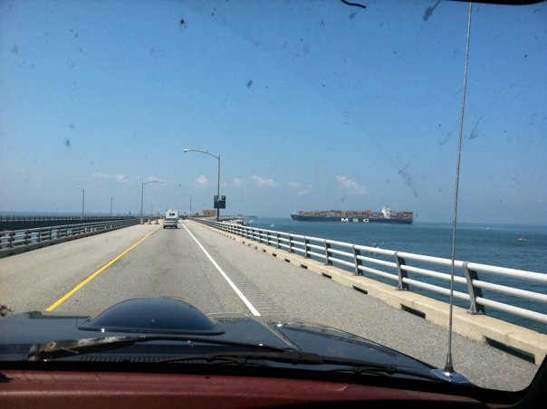 Dashboard of a red vehicle with a black hood scoop, seen from the interior looking out over a bridge with a cargo ship in the distance