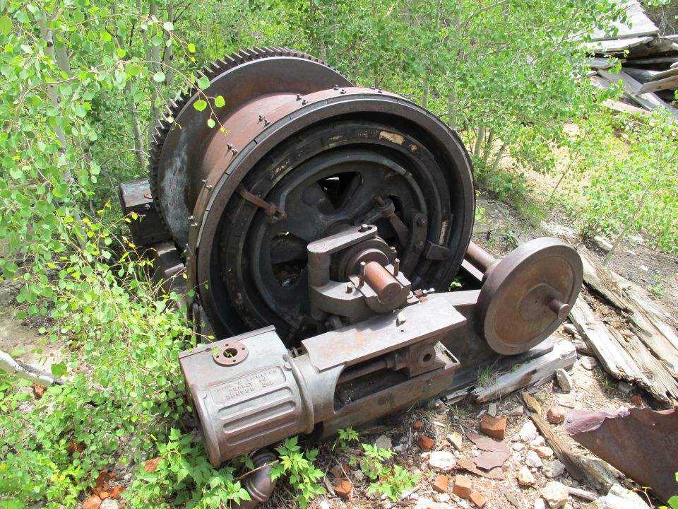 Large rusty industrial winch mechanism with a large gear and a smaller wheel, overgrown with vegetation