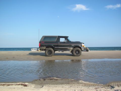 1995 Ford Bronco - A black Ford Bronco sits on a beach, its tires on the sand near the water's edge.