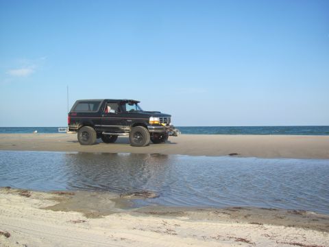 1995 Ford Bronco - A black 1995 Ford Bronco is parked on the sand near the water's edge on a sunny day.