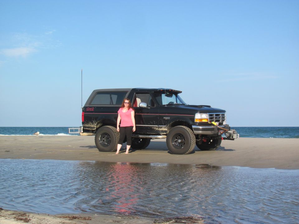1995 Ford Bronco on a sandy beach with the ocean in the background. Woman in pink shirt stands beside it