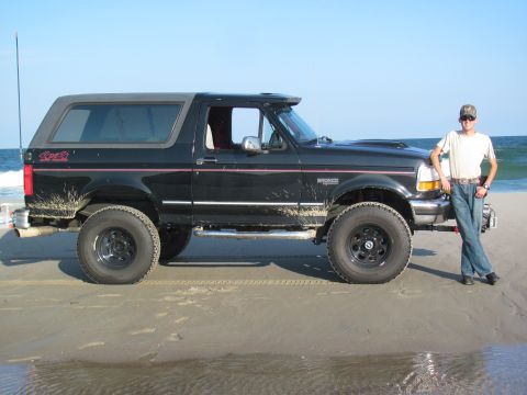 1995 Ford Bronco - A man poses next to a black Ford Bronco on a beach, with the ocean in the background.