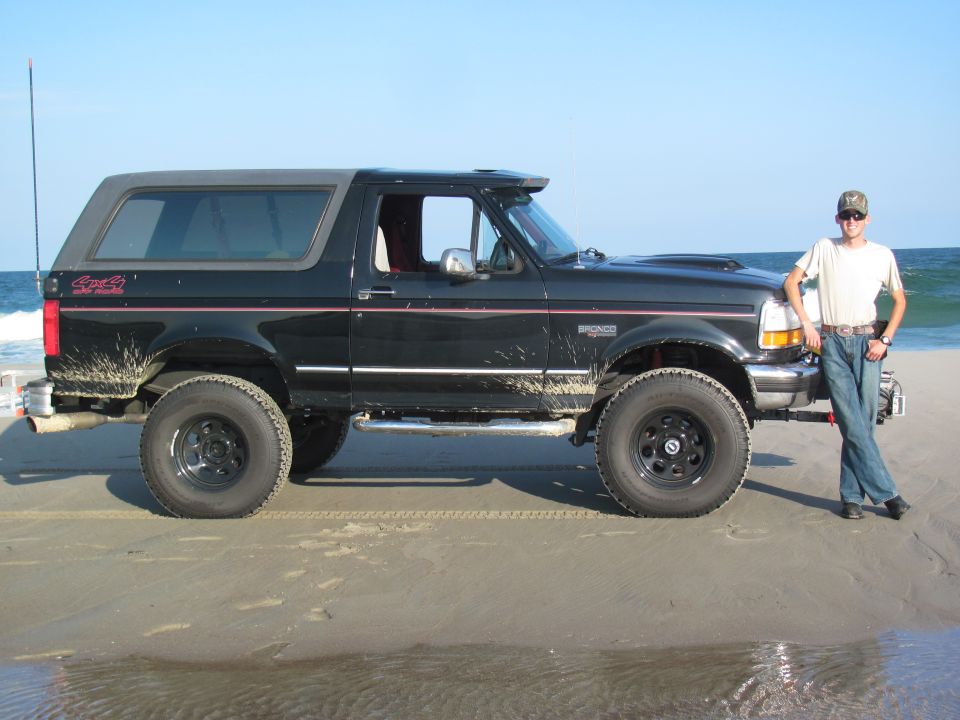 Black Ford Bronco on a sandy beach with a man standing by the front fender. The vehicle has mud splatters and a removable top