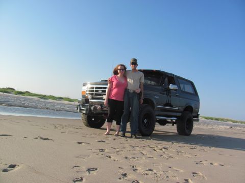 1995 Ford Bronco - Two people pose in front of a lifted Ford Bronco on a beach under a clear blue sky.