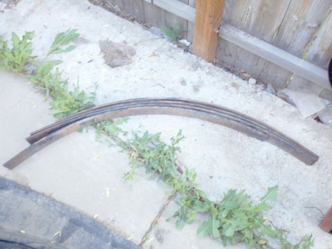 1989 Ford Bronco - Leaf spring part on concrete, with weeds and a wooden fence in the background.