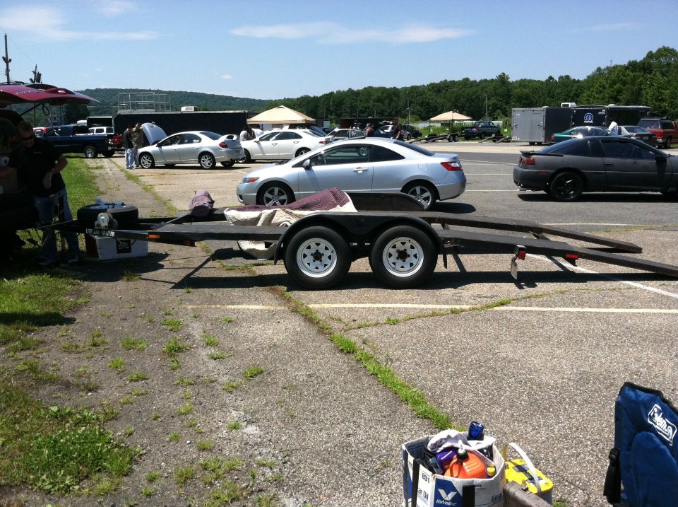 Cars and a trailer parked at a track. A silver coupe is in the foreground, with a dark sedan and other vehicles in the background