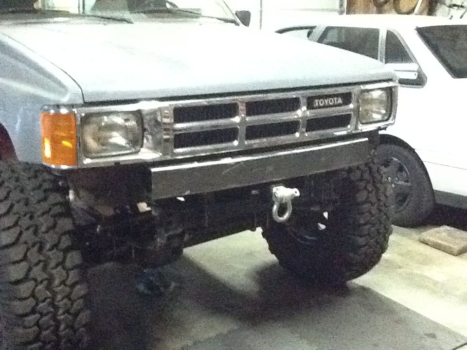 Toyota truck front end with aftermarket bumper and large tires. Another white vehicle is visible behind it