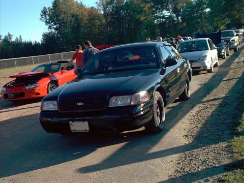 2000 Ford Crown Victoria - A line of cars, including a black 2000 Ford Crown Victoria, are parked on a dirt road at an outdoor event.