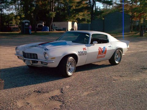 2000 Ford Crown Victoria - A white 1970s Chevrolet Camaro with racing decals is parked on a gravel lot.