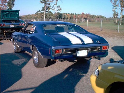 2000 Ford Crown Victoria - A blue muscle car with racing stripes and an SS badge is parked next to a yellow car, with another car on a trailer in the background.