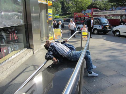 1968 Ford Mustang - A man in a striped shirt and jeans lies reclined across a metal handrail on a city sidewalk, with red double-decker Edinburgh sightseeing t…