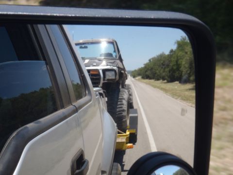 1979 Ford Bronco - A heavily modified off-road vehicle with large tires is seen being towed on a highway, reflected in the side mirror of another vehicle.