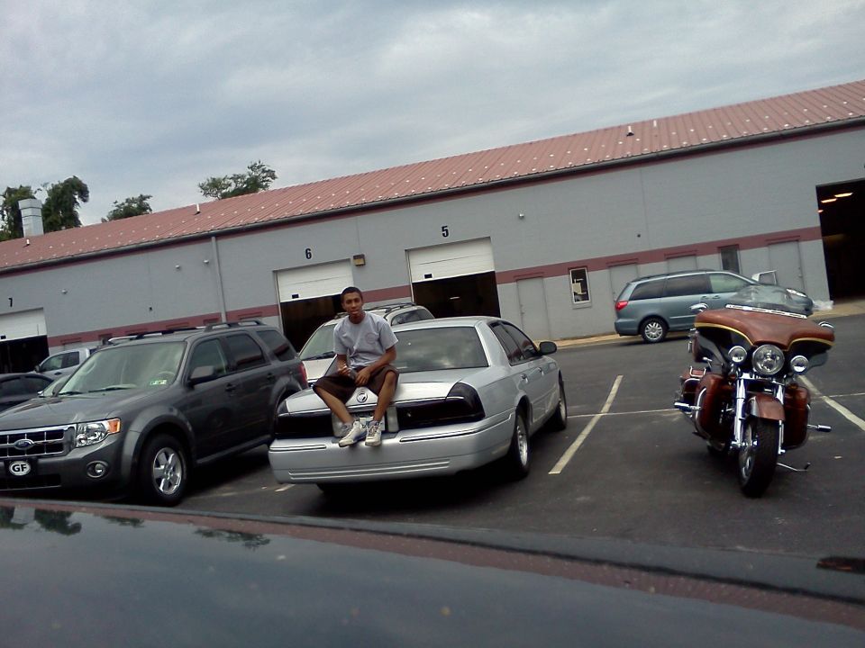 Man sitting on the trunk of a silver sedan parked in a lot. Another SUV and a motorcycle are also visible