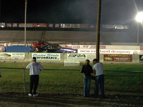 2000 Ford Crown Victoria - Two cars race down the drag strip at New England Dragway under the lights, observed by spectators.