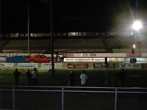 2000 Ford Crown Victoria - A red sedan is visible at the New England Dragway, with spectators in the foreground and stadium seating in the background.