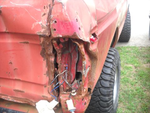 1993 Ford F-150 - Close-up of a damaged red truck's rear quarter panel, showing rust and exposed wiring where the taillight should be.