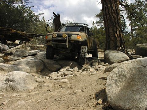 1997 Ford Ranger - Mud-covered tan Ford Ranger pickup crawling over granite boulders on a pine-forested mountain trail, front three-quarter view showing a tub…
