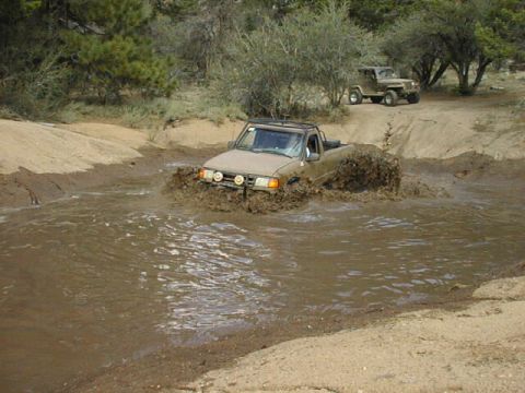 1997 Ford Ranger - A tan compact pickup with a roll bar and round driving lights on the front bumper blasts through a muddy water crossing, throwing a brown w…