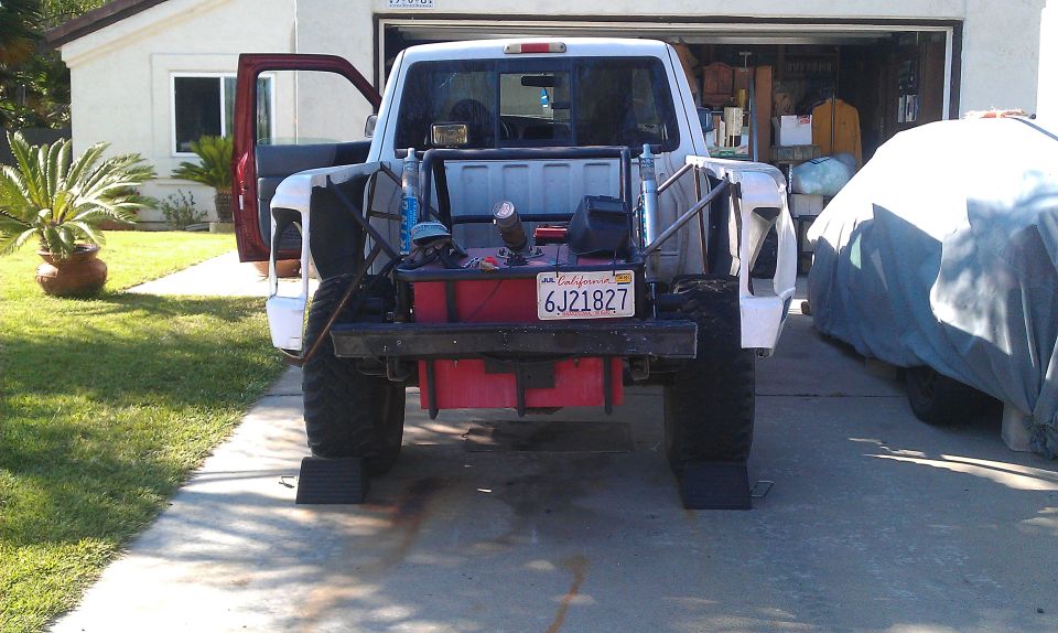 Rear view of a white truck with a custom bed cage, fuel tank, and off-road tires. Another vehicle is covered on the right