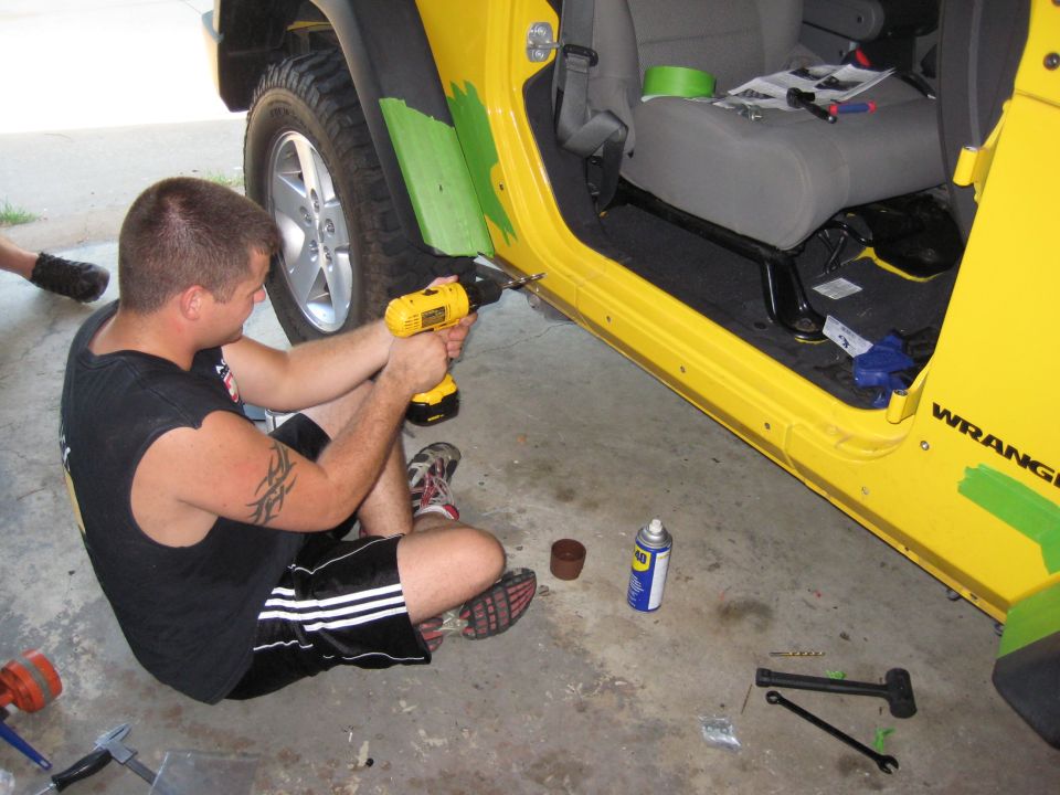 Man drilling into the door sill of a yellow 2005 Jeep Wrangler. Tools and a can of lubricant are on the ground