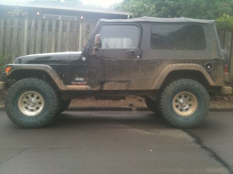 2005 Jeep Wrangler - A muddy black Jeep Wrangler Unlimited is parked on a street, showing its side profile and large off-road tires.