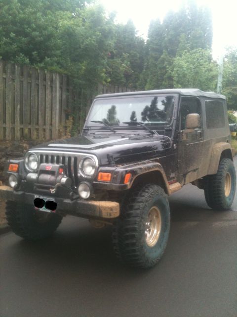 2005 Jeep Wrangler - A black 2005 Jeep Wrangler with a winch and off-road tires parked on a wet street.