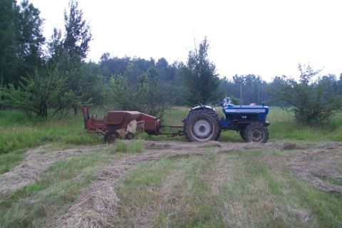 2000 Chevrolet Tahoe - A blue tractor tows a red baler through a field of cut grass under an overcast sky.
