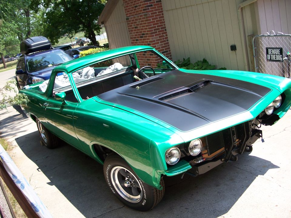 Green 1970 Ford Ranchero with black hood stripes, missing front bumper and grille, parked on a driveway