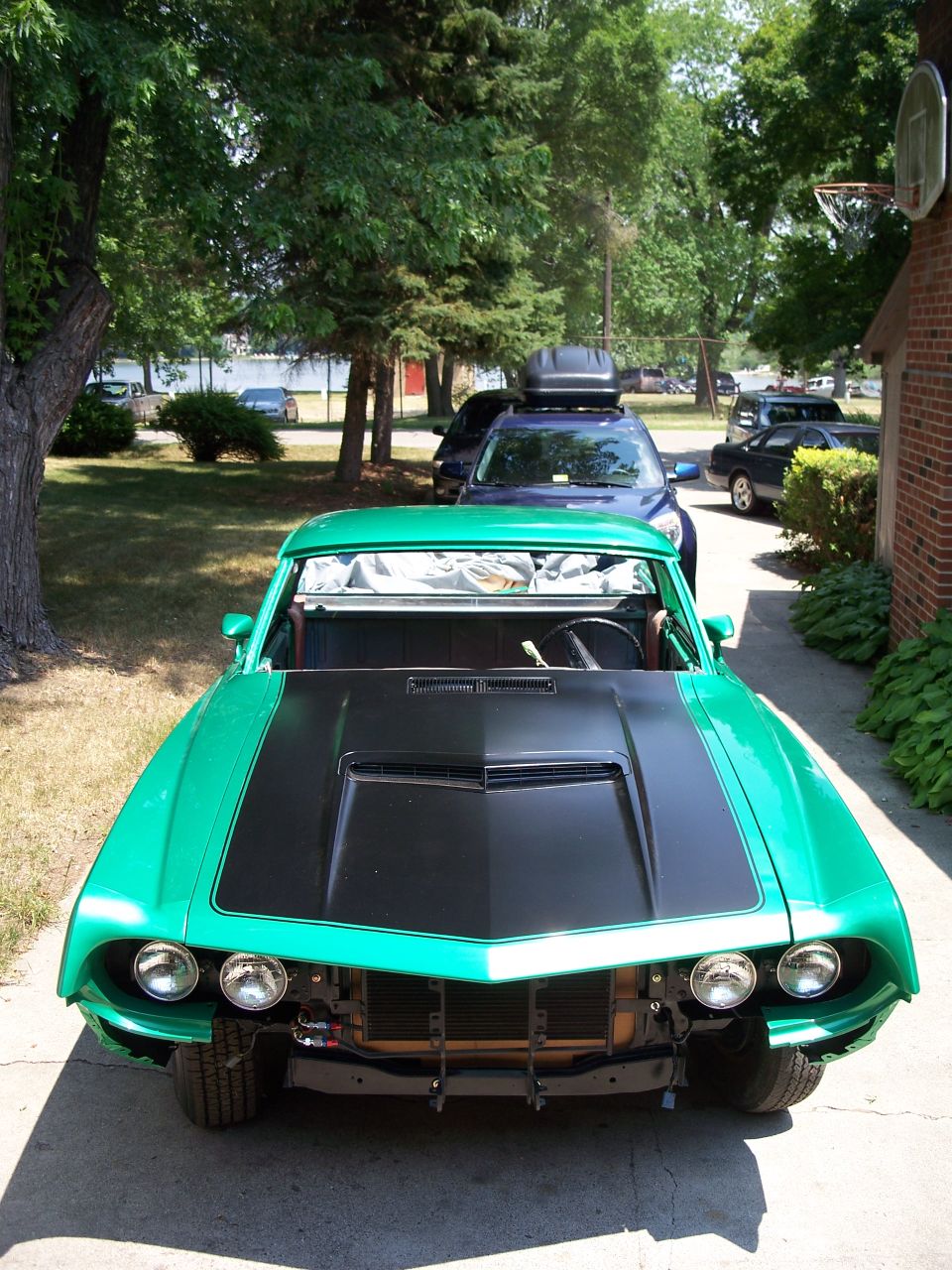Green 1960s muscle coupe head-on in driveway, grille and windshield removed, black cowl hood