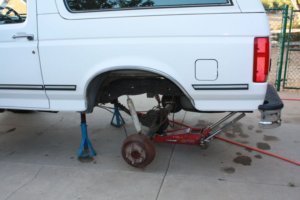 White Ford Bronco on jack stands, rear wheel removed showing drum brake hub, red floor jack under axle