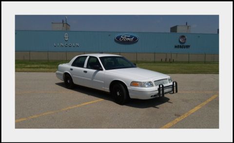 1998 Ford Crown Victoria - A white 1998 Ford Crown Victoria, equipped with a push bar, is seen parked in front of a building displaying Ford, Lincoln, and Mercury bra…