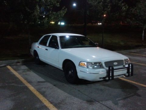 1998 Ford Crown Victoria - A white sedan, likely a Ford Crown Victoria, is parked in a lot at night, equipped with a push bumper.