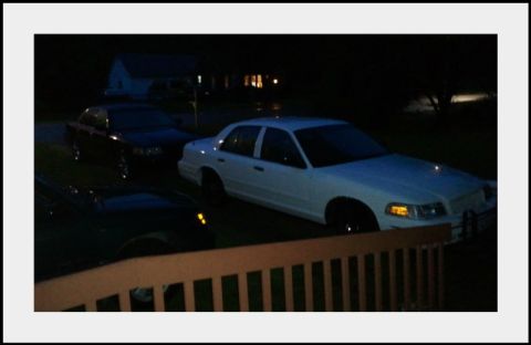 1998 Ford Crown Victoria - Elevated night view over a wooden deck railing of a white Ford Crown Victoria parked in a driveway with its amber parking lights glowing an…
