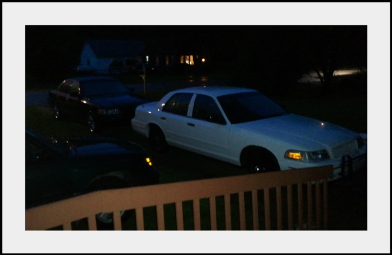 White Ford Crown Victoria in driveway at night with parking lights on, seen over deck railing
