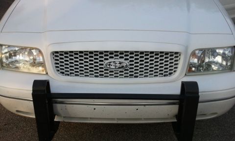 1998 Ford Crown Victoria - Close head-on view of a white Ford's front end showing a chrome honeycomb mesh grille with the blue Ford oval badge centered in it. Clear h…