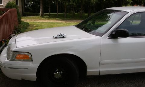 1998 Ford Crown Victoria - Low front three-quarter shot of a white Ford Crown Victoria in a grassy driveway, with a small white die-cast police cruiser model posed on…