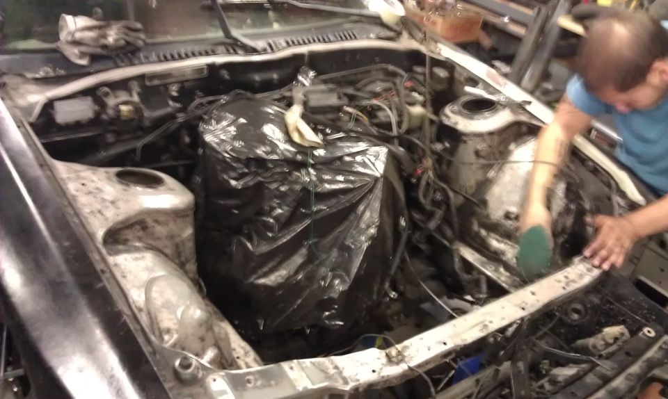 1987 Mazda RX-7 engine bay with engine covered in a black plastic bag, being cleaned by a person