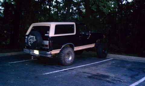 1990 Ford Bronco - Lifted black and tan Ford Bronco parked in a lot at night, showing its rear profile and spare tire.