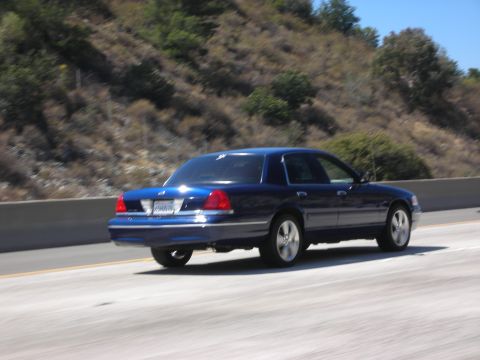 2004 Ford Crown Victoria - A blue Ford Crown Victoria is seen from the rear on a highway with a dry, grassy hillside in the background.