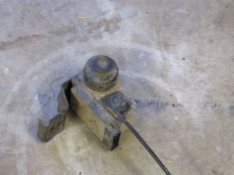 1995 Ford Bronco - A worn metal component with a cable, likely a vehicle part, sits on a concrete surface.
