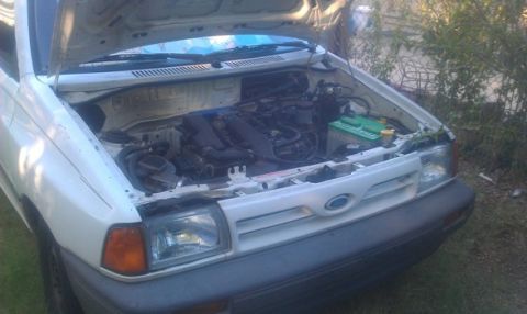 1991 Ford Festiva - The front of a white hatchback is shown with its hood raised, revealing the engine compartment and a green battery.