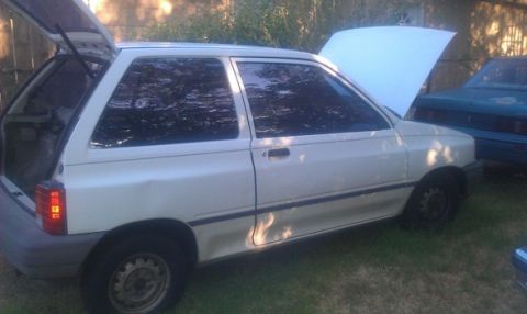 1991 Ford Festiva - A white 1991 Ford Festiva hatchback is parked outside with its rear hatch and hood open.