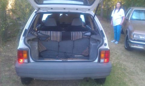 1991 Ford Festiva - The rear of a white hatchback is shown with its trunk open, revealing interior fabric. Another vehicle is visible to the right.