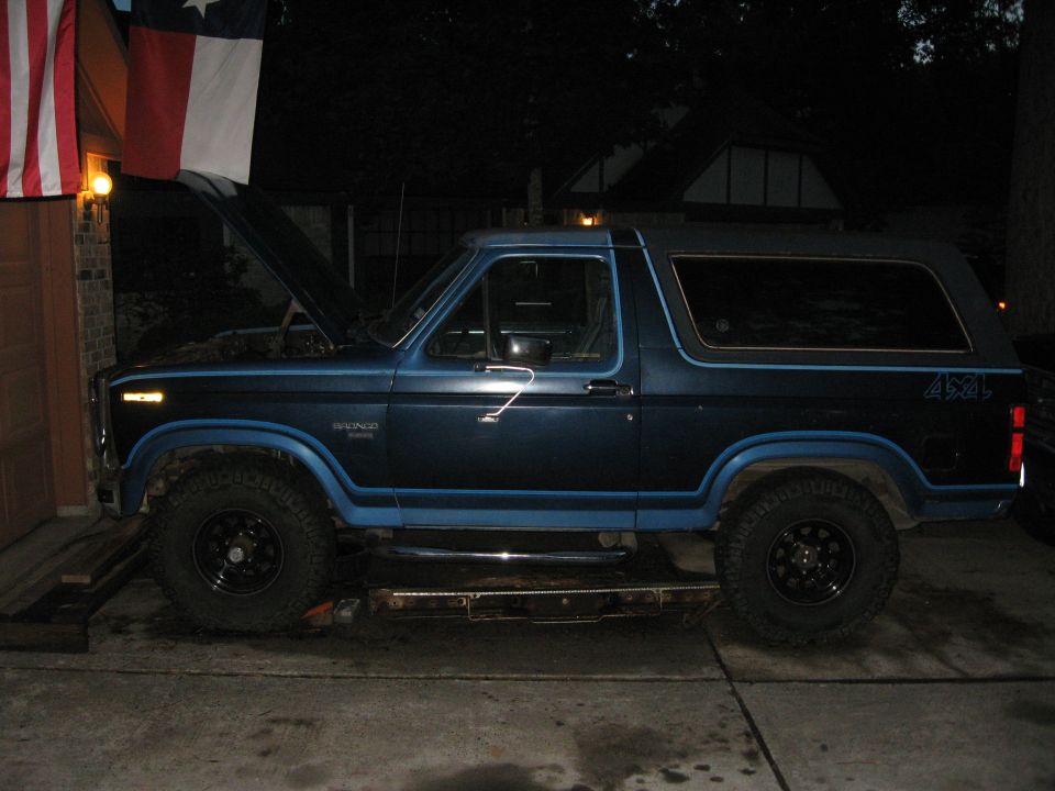 Dark blue Ford Bronco side view at night with hood open, parked on ramps in a driveway near hanging flags