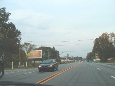 1997 Ford Taurus - dark-colored sedan travels down a two-lane rural highway lined with billboards and roadside businesses under a gray sky.