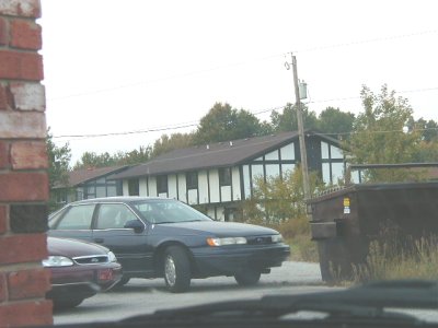 1997 Ford Taurus - Shot through a windshield past a brick pillar: a dark blue third-generation Ford Taurus sedan parked on pavement with a red car behind it,…