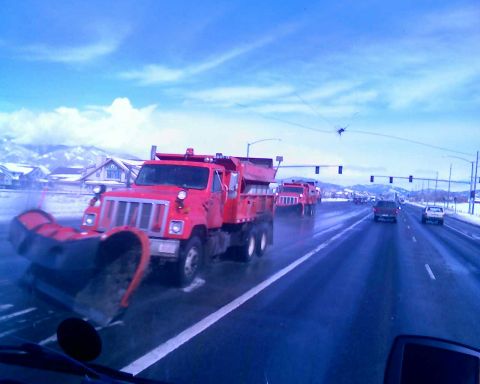 2006 Ford E350 - Multiple red snow plow trucks drive down a highway on a cloudy day with snow-covered mountains in the background.