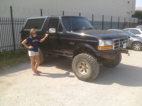 1995 Ford Bronco - A black 1995 Ford Bronco is shown with a woman standing beside it, highlighting its lifted suspension and off-road tires.