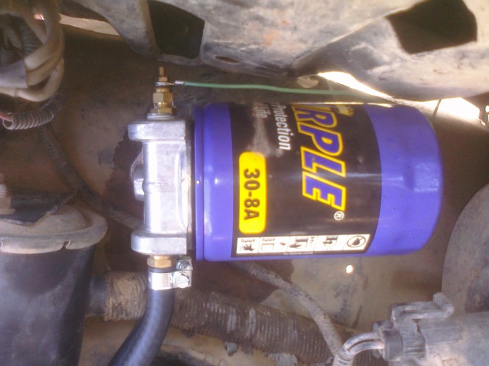Purple fuel filter with yellow label showing 'TRIPLE 30-8A' and diagrams, connected to metal fittings and black hose